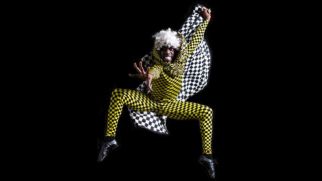 a dancer portraying the mouse king jumps up and reaches one hand out toward the camera. He is wearing a yellow and black checkered suit and is holding a checkered flag above his head. He looks intensely mischievous.